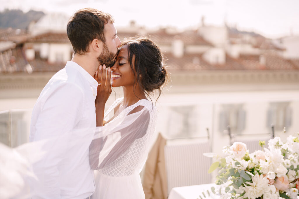 Destination wedding in Florence, Italy. Caucasian groom and African-American bride cuddle on the roof, in the sunny sunset light.